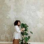 A woman in a white shirt tends to a monstera plant against a concrete wall, reflecting a minimalist interior design.