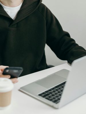 Casually dressed man working from home using a laptop and smartphone, with a coffee cup nearby.