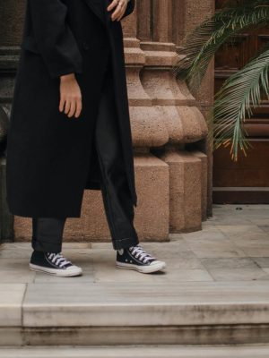Crop view of unrecognizable person in black trousers and coat with sneakers standing on porch of stone building on daytime